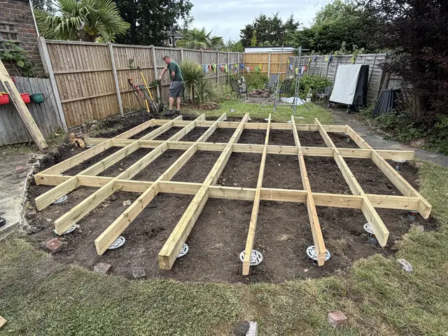 Timber floor grid being assembled over visible ground screw heads in an enclosed garden.