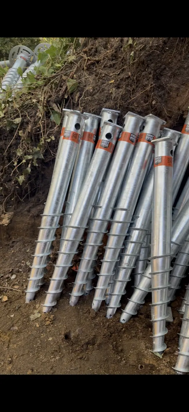Several galvanised helical ground screws lying together on soil and vegetation.