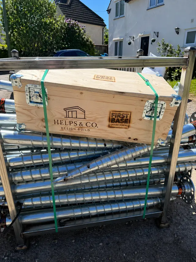 A strapped timber transport crate marked Helps and Co and First Base Ground Screws beside galvanised screw components.