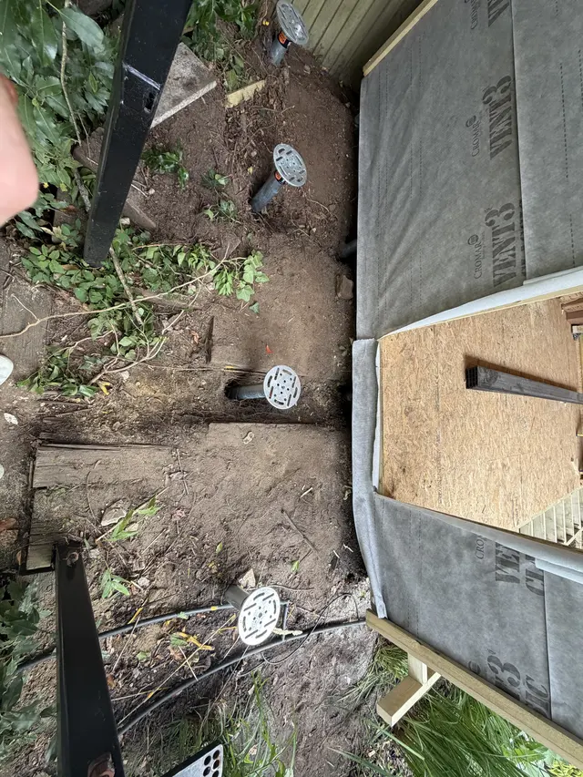 Several installed ground screw heads beside a building under construction and close to garden vegetation.